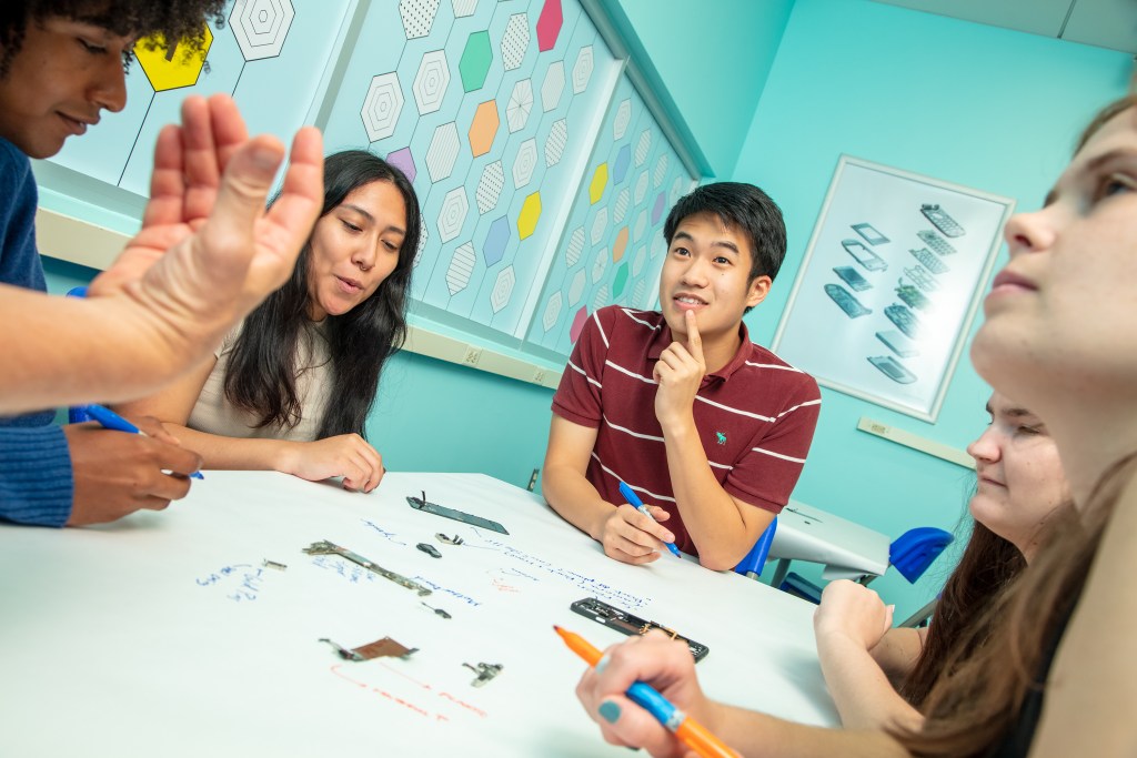 A diverse group of teenagers sits around a paper-covered table where several cellphones have been taken apart. The students use markers to make notes on the paper about the cellphone components.