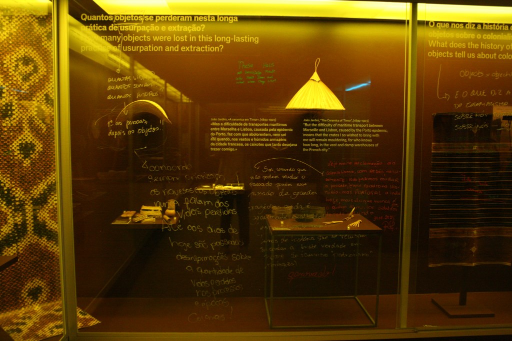 A glass-fronted floor-to-ceiling display case holds a collection of Timorese fibre-woven hats and combs against a dark brown backing. Words and arrows in white, green, and red marker are hand-written across the glass, referring to the printed text and other visitors’ comments.