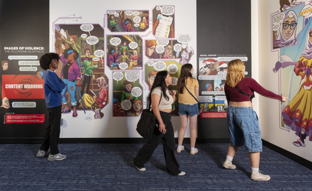A diverse group of teenagers looks at a museum gallery wall covered with a comic-book-style mural.