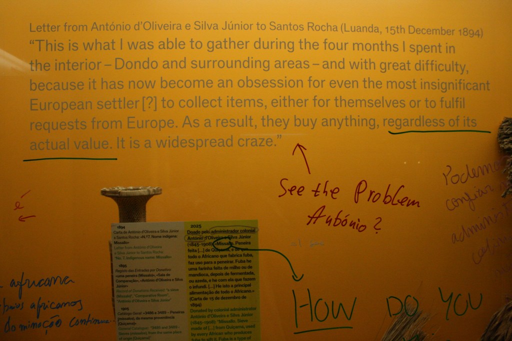 A close-up of a text by António d’Oliveira, placed in front of an Angolan vase against a warm-yellow backing. Hand-written red, green, and white commentaries respond to and critique the labels’ contents.