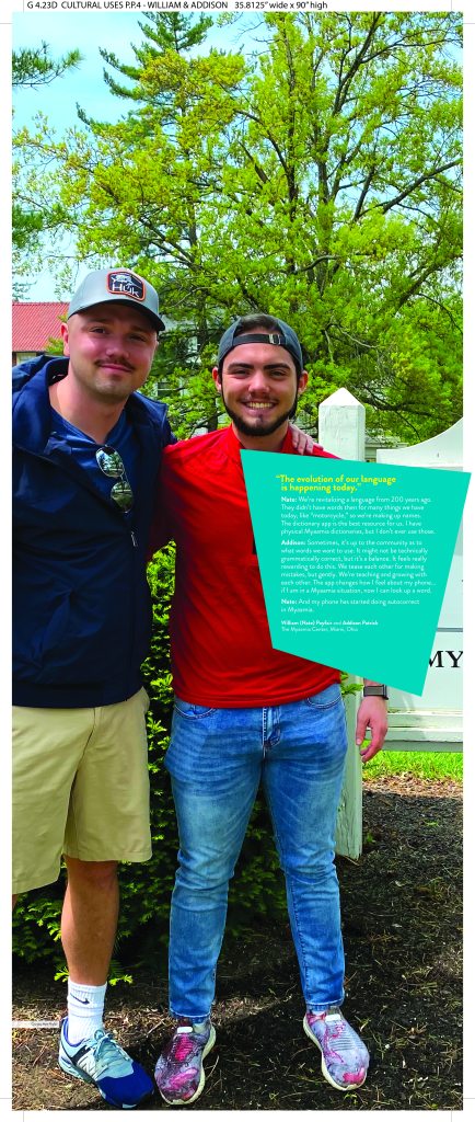 A museum exhibition panel includes text and a large photo of William (Nate) Poyfair and Addison Patrick, two young men wearing casual clothes with their arms draped over each other’s shoulders.