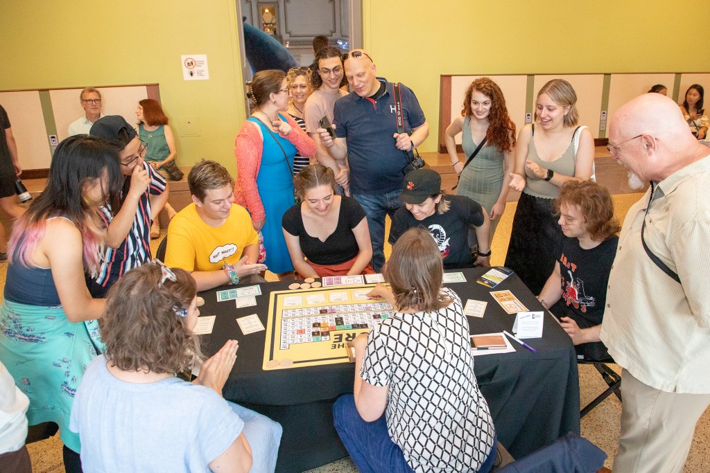 A diverse group of 14 museum visitors surrounds a table where a museum employee facilitates a board game.