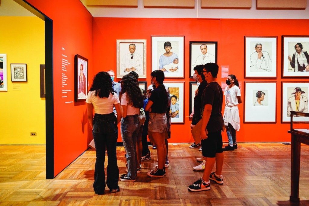 Group of students standing in front of a red gallery wall with portraits displayed on it. 