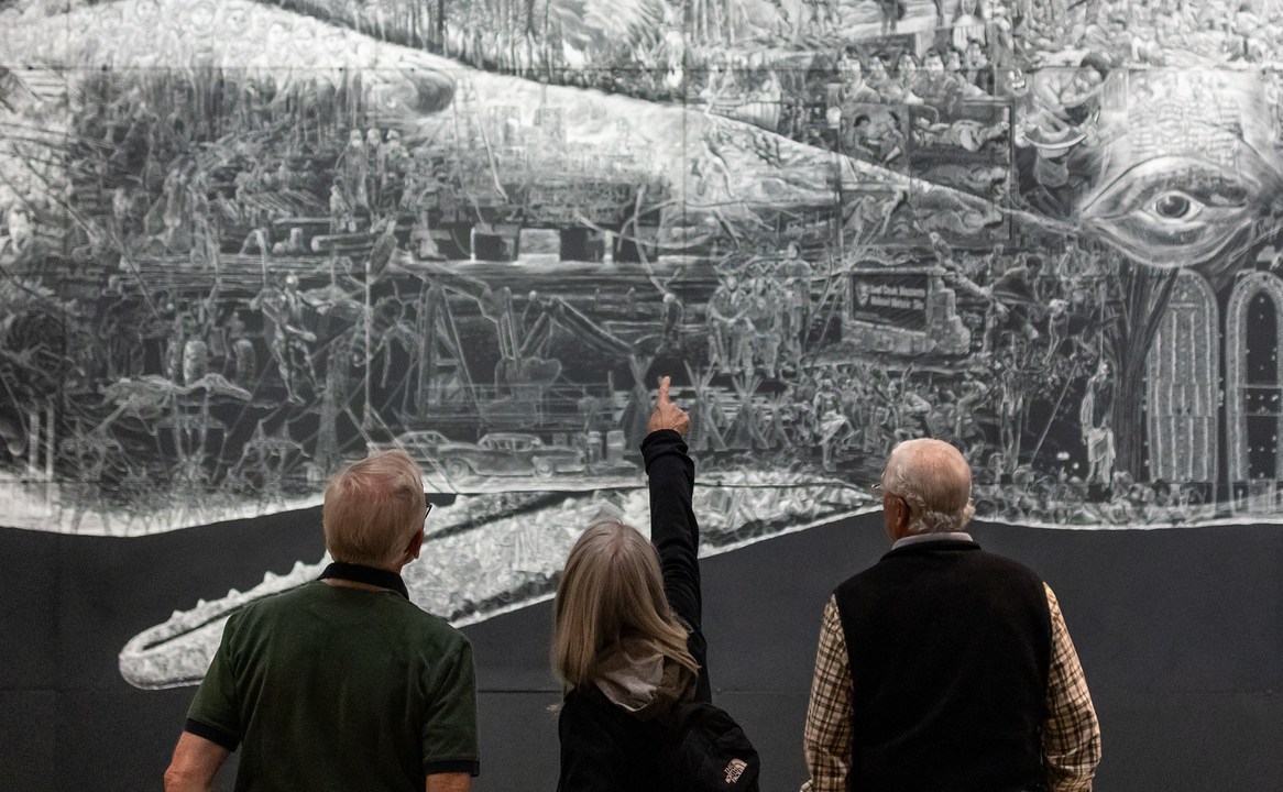 Two older white men with short white hair and one older white woman with shoulder-length white hair face the mural Or, the Whale. The woman points upward toward an illustration on the mural, and the men look to where she is pointing.