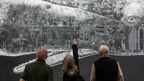 Two older white men with short white hair and one older white woman with shoulder-length white hair face the mural Or, the Whale. The woman points upward toward an illustration on the mural, and the men look to where she is pointing.