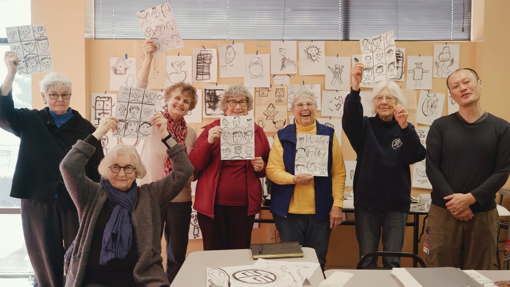 Group of older women and one man holding up drawings of faces.