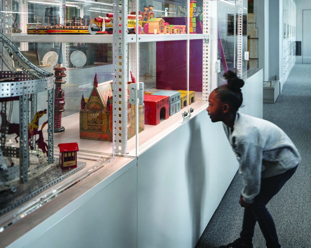 A young girl is looking at vintage toys in a display case in an open-storage gallery of a museum.