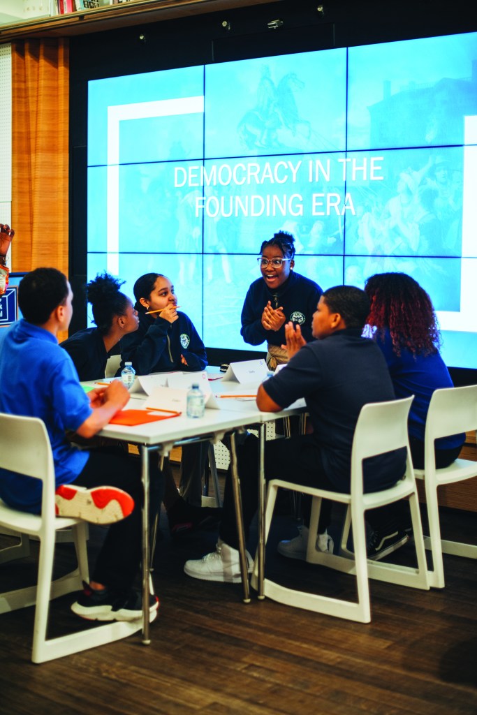 Group of students seated at a table together, having a discussion in front of a screen that says: "Democracy in the Founding Era"