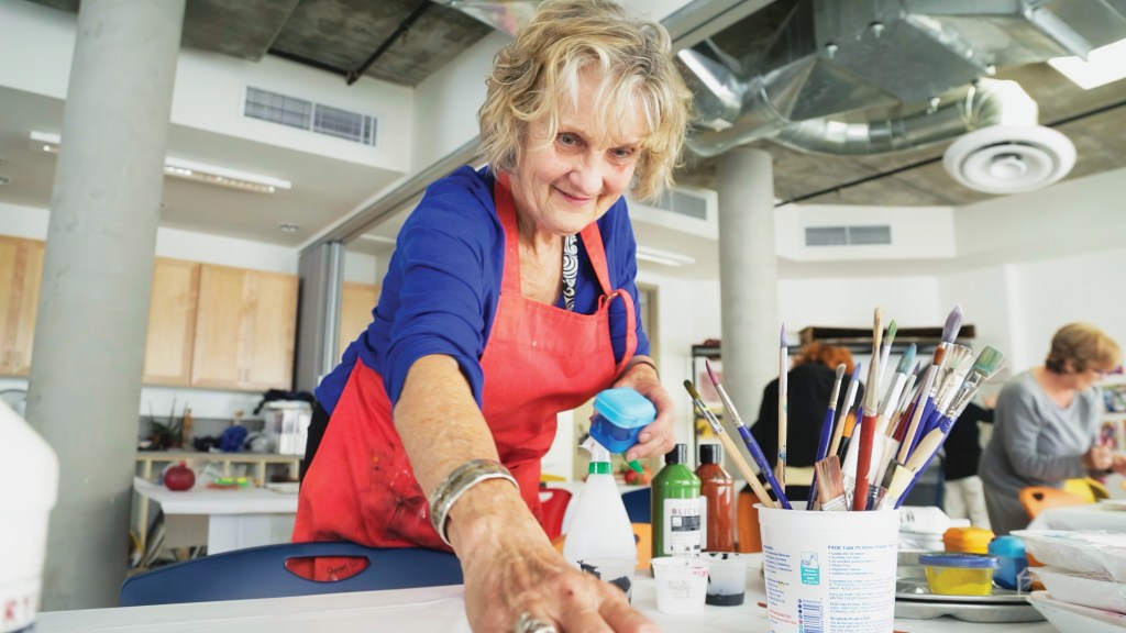 Older woman wearing a red apron doing an art project in as museum with paintbrushes on a table.