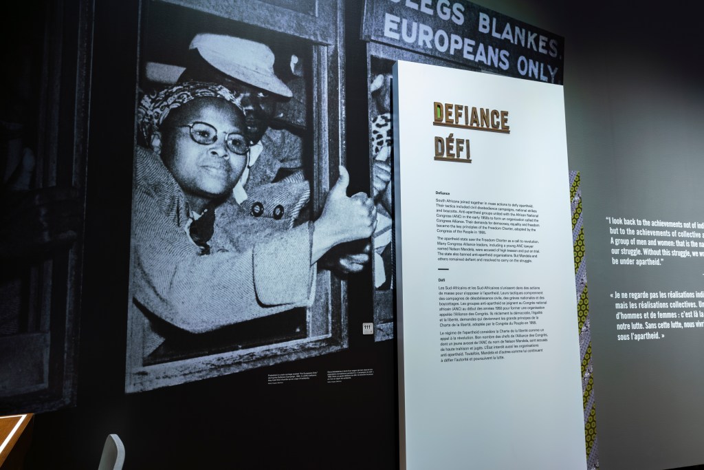 A large black-and-white photograph covers the left side of the exhibition wall. It shows a Black woman leaning out of the window of a crowded train. She holds her arm out with her thumb raised. Above the window, a sign reads “EUROPEANS ONLY” in English and Afrikaans. To the right, a vertical exhibition panel titled “Defiance / Défi” presents bilingual text. Further to the right, part of another panel is visible.