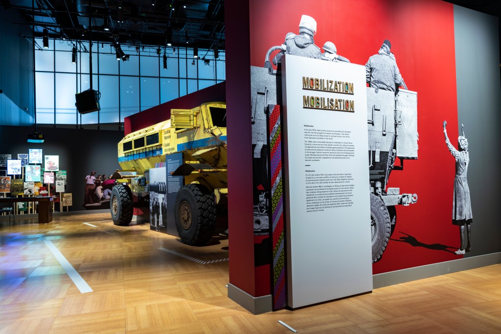 A large yellow armored vehicle is displayed in the center of a gallery space. To the right, a tall interpretive panel titled “Mobilization / Mobilisation” stands in front of a red wall featuring a black-and-white mural of soldiers riding on the back of a military vehicle. A cutout figure of a woman with one arm raised appears beside them. In the background, additional exhibit elements include posters, photographs, and display cases.