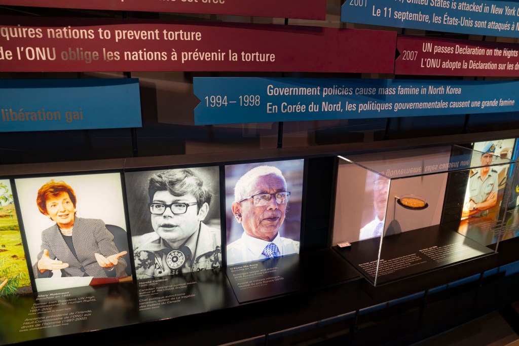 A row of illuminated portrait panels displays several individuals associated with human rights work. To the right, a small object is presented in a clear display case. Above the portraits, suspended timeline panels in English and French highlight major global human rights events.