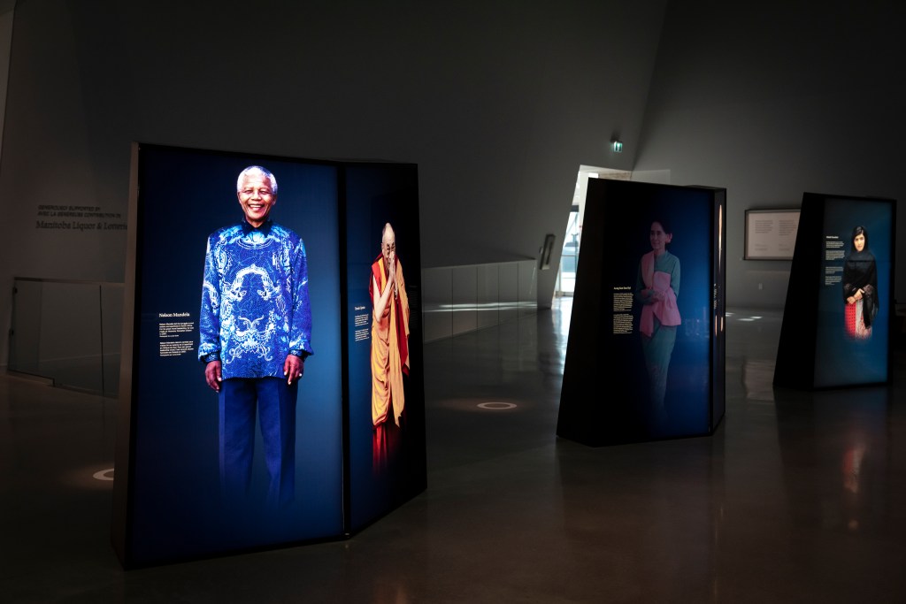 Illuminated portrait panels stand throughout a dim gallery space. The nearest panel shows a person smiling and wearing a patterned shirt; beside it is a portrait of a person in traditional robes with hands held together. Farther back, two additional portrait panels show individuals standing against dark backgrounds. The third portrait from the left is dimmed.