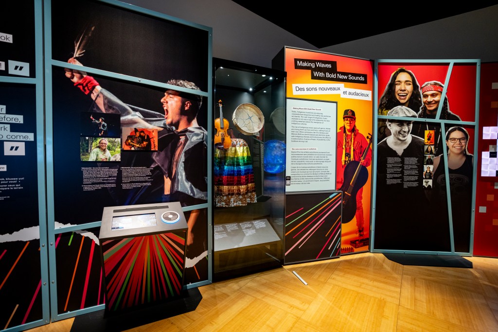 A colourful exhibition section features large photographic panels of musicians and performers. At the center, a glass case displays a drum, a fiddle, and a brightly striped skirt. To the right, a panel titled “Making Waves With Bold New Sounds / Des sons nouveaux et audacieux” stands beside portraits of several smiling individuals. An interactive station with multicoloured stripes appears in the foreground.