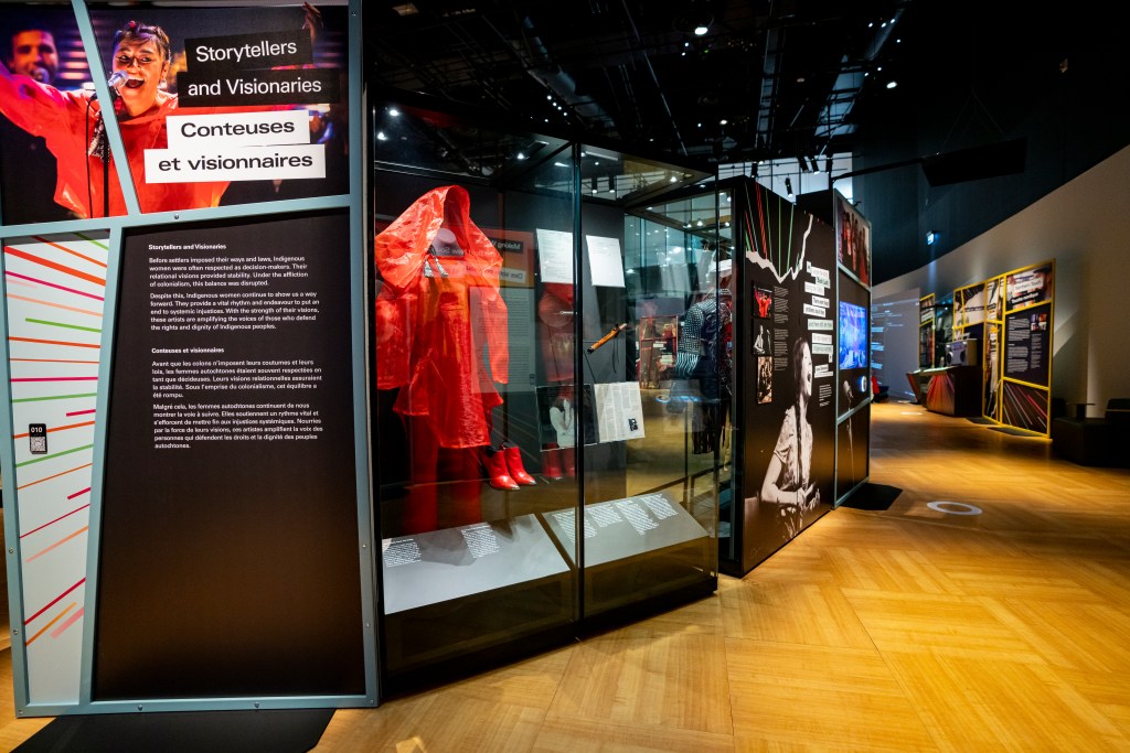 An exhibition section titled “Storytellers and Visionaries / Conteuses et visionnaires” features a large text panel beside a glass display case containing a bright red coat and matching boots. Additional objects and documents are visible further along the case. To the right, another panel displays photographs and text, with more exhibit installations extending into the background of the gallery.