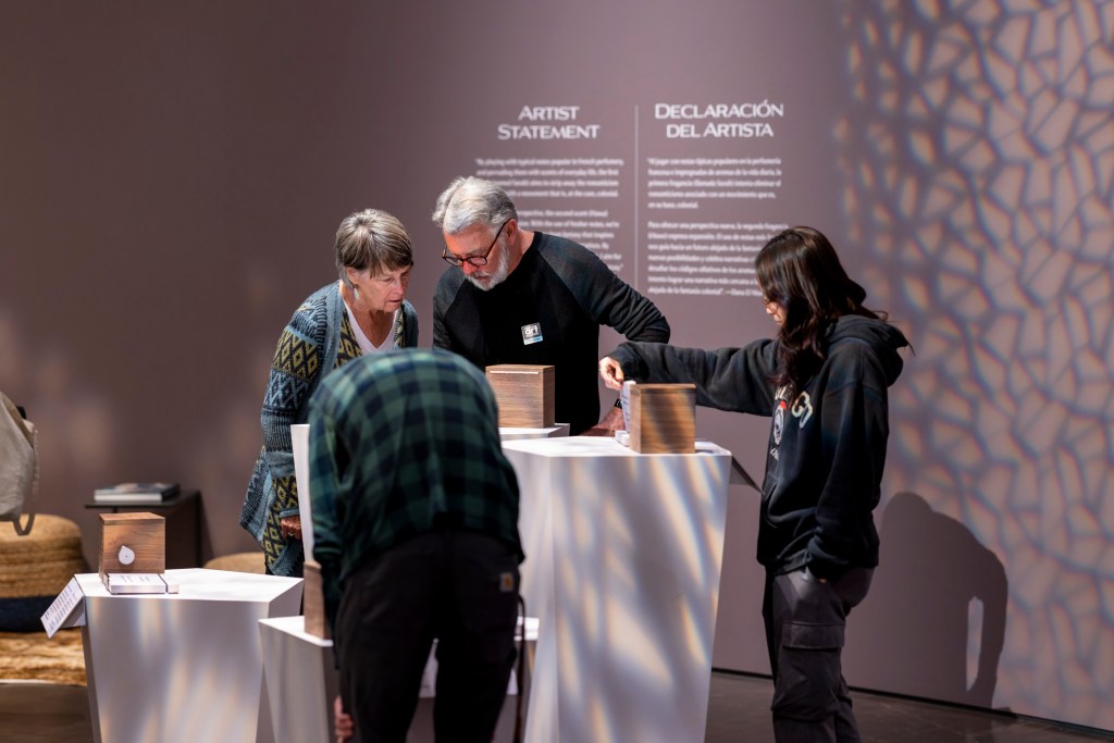 Four museum visitors gathered around individual scent boxes on pedestals in various stages of engagement. A couple reads a label; a young woman lifts a flap to look at the scent ingredients; and a man bends over to smell inside a box.