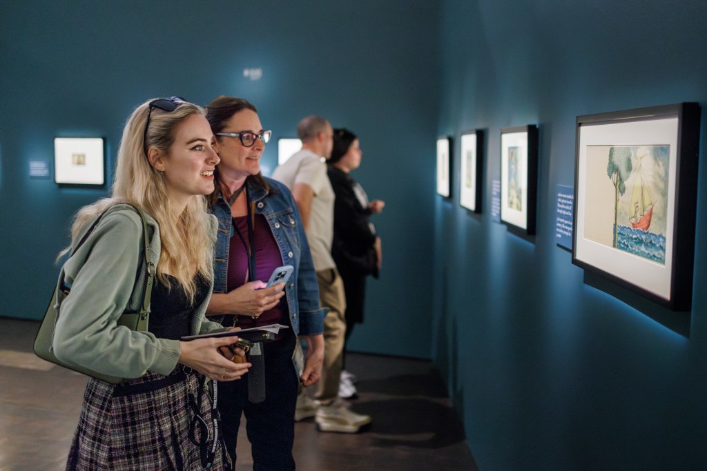 Two women, on the left, smiling and looking at, on the right, a framed picture hanging on the wall from Maurice Sendak’s book Where the Wild Things Are. On the wall to the left of the framed picture is a label with text that accompanies that picture in the book. In the background, more framed pictures with book text to the left of them are on the wall, and two additional blurred people are looking at one of those pictures.