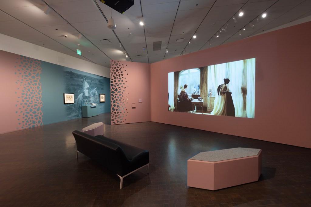 ] A three-paneled wall in an art exhibition shows a projected film still of two animated women in an imagined interior. An empty bench is situated in front of the projected film. In the background, another section of the gallery is visible with paintings and wall graphics. 