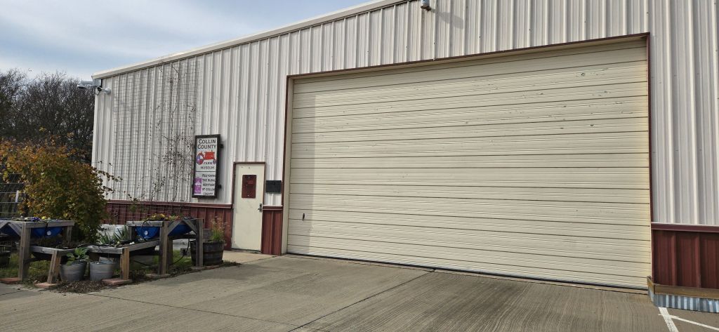 A large metal-sided building with a tall, closed beige roll-up garage door and a smaller single door beside it. A sign next to the door reads โCollin County Farm Museum,โ with additional text about preserving agricultural heritage. Several outdoor planter boxes with plants sit to the left of the entrance, and security cameras are mounted near the roofline. A concrete driveway extends across the foreground.