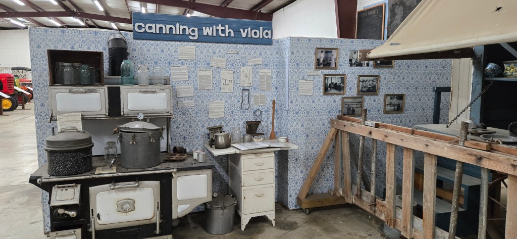 Exhibit titled โCanning with Violaโ showing a recreated historic farm kitchen inside a large warehouse space. The display includes an early 20th-century wood-burning stove with canning jars, pressure canners, and cookware arranged on and around it. Blue-patterned wallpaper lines the modular exhibit walls, which feature interpretive text panels, diagrams, and black-and-white photographs documenting home canning practices. A small worktable holds kitchen tools and jars, while a wooden rack and suspended scale appear on the right, illustrating food preparation and preservation.