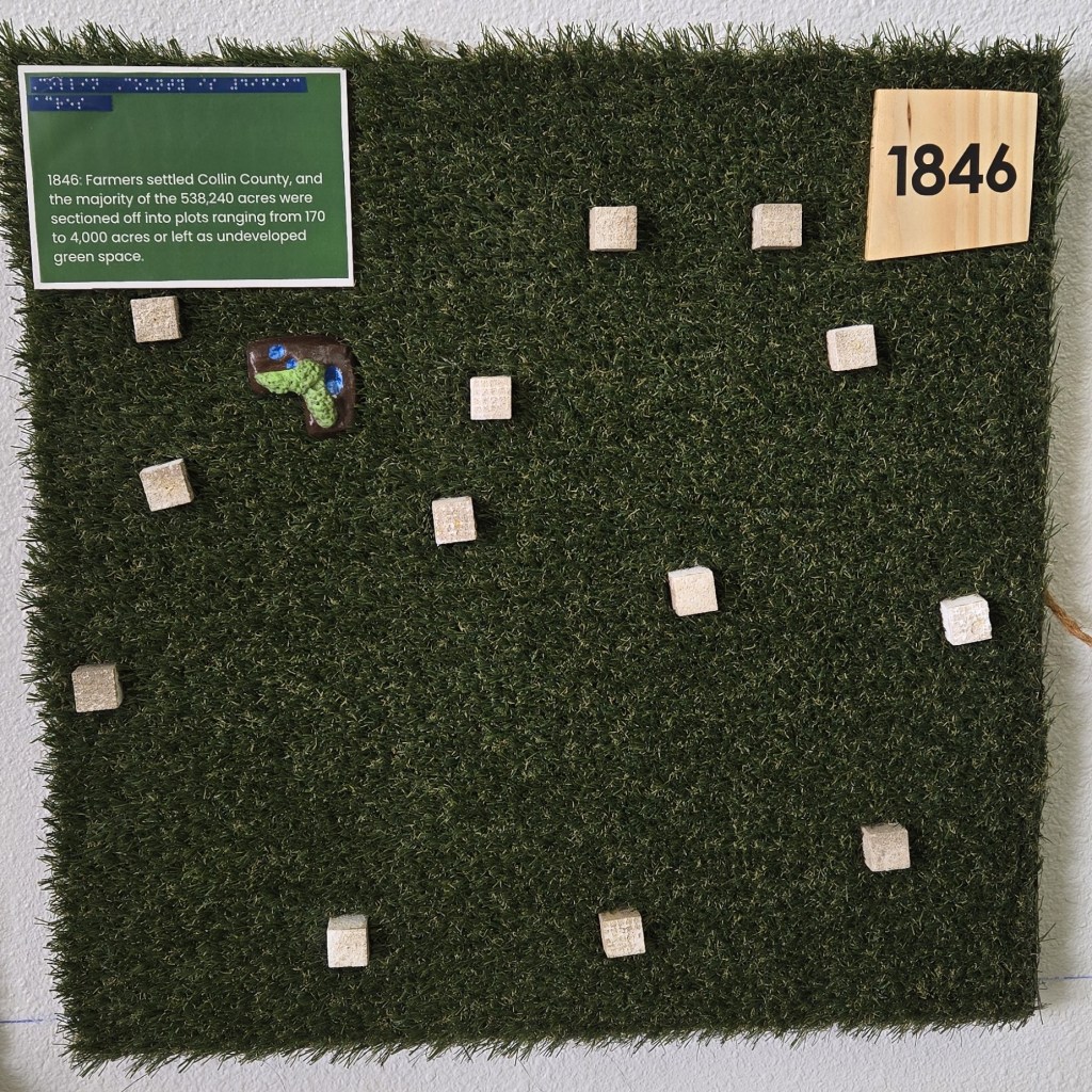 ] A square panel covered in artificial grass features small beige cube markers arranged across its surface, along with a single green-and-blue L-shape of Myers Park & Event Center. A wooden plaque in the upper right corner reads “1846.” A green sign with braille and printed text explains that in 1846, farmers settled Collin County and divided its 538,240 acres into plots ranging from 170 to 4,000 acres or left the land as undeveloped green space.