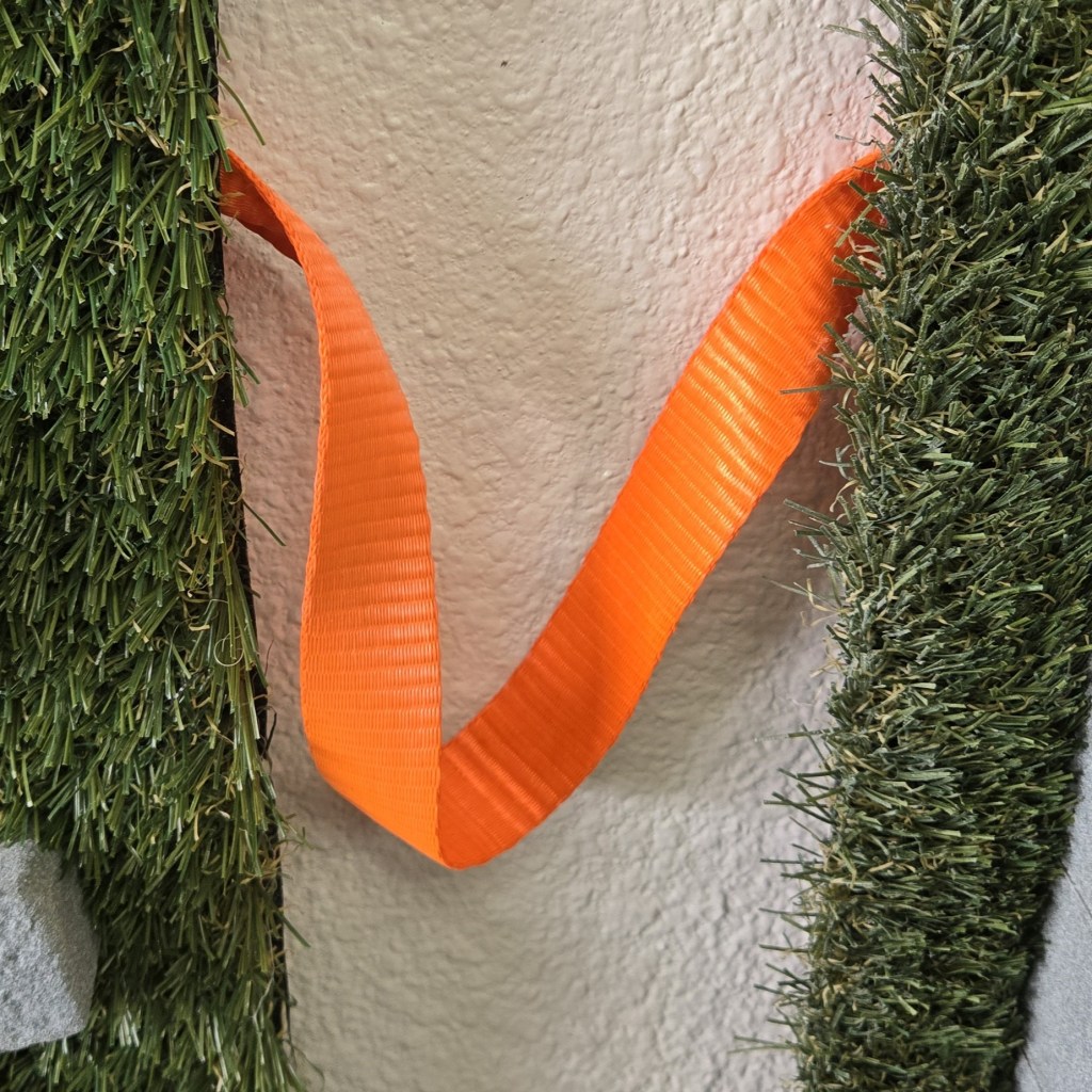 A close-up view of a bright orange nylon strap hanging loosely in a curved โVโ shape against a textured white wall. On both sides of the strap are panels covered in artificial green grass, creating a narrow vertical gap where the strap is suspended.