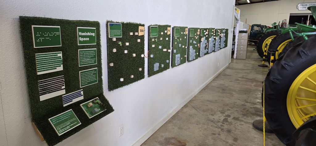 A long wall inside the Collin County Farm Museum displays a series of tactile green panels covered in artificial grass. The panels feature raised blocks, braille labels, printed text, and small landscape models. The first panel, titled โVanishing Space,โ includes several braille and text sections explaining the exhibitโs theme. Additional panels show different years, including โ1846,โ with increasing numbers of blocks representing development. To the right, a row of large John Deere tractors with bright yellow wheels is visible, creating an aisle.