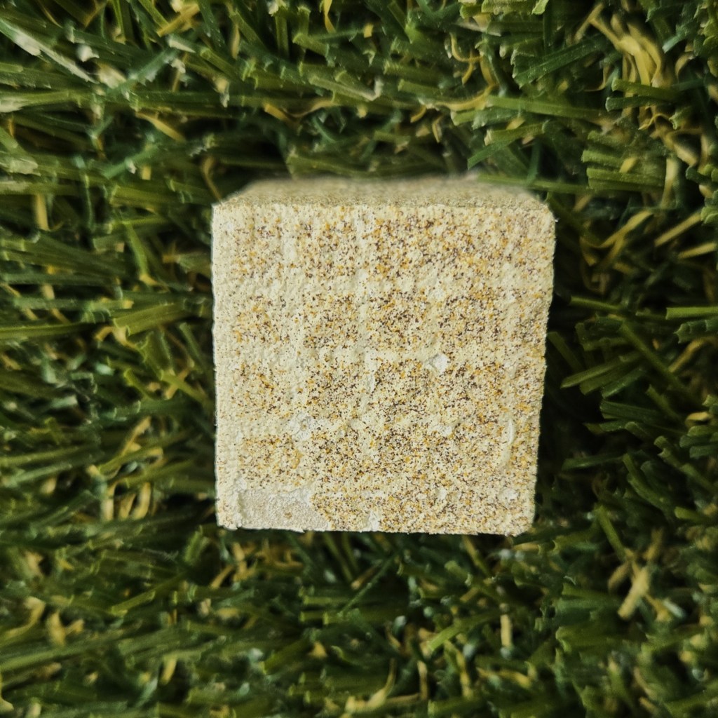 A close-up view of a small textured beige cube resting on a background of artificial green grass. The cube has a rough, speckled surface resembling sandstone with a faint grid of white lines.