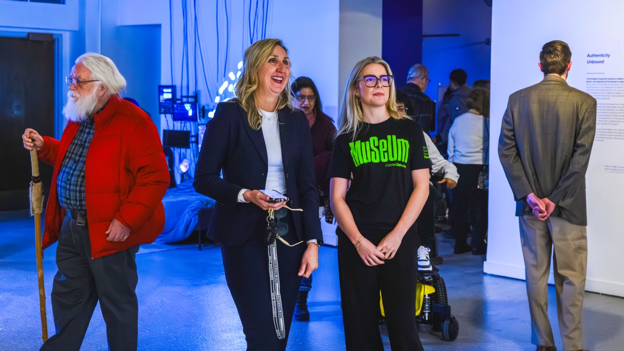 Visitors walk through a gallery during an opening reception, with one attendee engaging in conversation with a Museum CoLaborator while viewing the installation Dark Loops in the Blurred Realities exhibition.