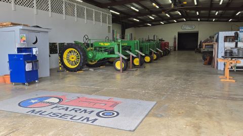 Interior view of Collin County Farm Museum featuring a row of restored John Deere tractors lined up side by side. A gray floor mat with the Museum logo sits in the foreground. To the right are displays of vintage kitchen and canning equipment, and to the left is a small, shed-like structure and a blue utility cart. The space features a high metal ceiling, bright overhead lights, and concrete flooring.