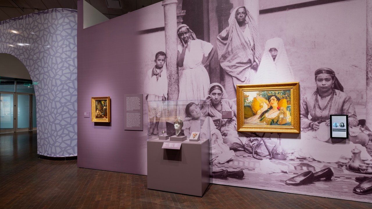 A wall inside an art exhibition with two oil paintings of women lounging and a small jewelry case accompanied by object labels and a wall text panel. Behind the case and one of the paintings is a large graphic of a 19th-century photograph of Algerian women and their children. A small touch screen is on the far right of the second painting.