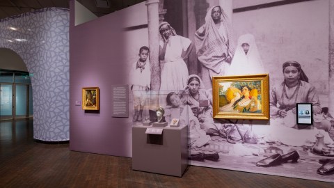 A wall inside an art exhibition with two oil paintings of women lounging and a small jewelry case accompanied by object labels and a wall text panel. Behind the case and one of the paintings is a large graphic of a 19th-century photograph of Algerian women and their children. A small touch screen is on the far right of the second painting.