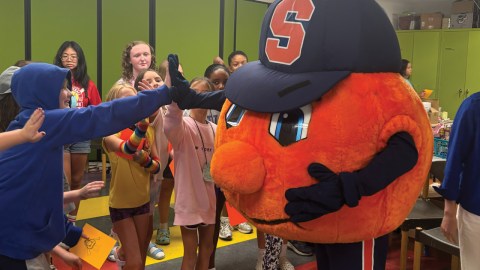 Orange Syracuse University mascot greets group of children in a room with green walls