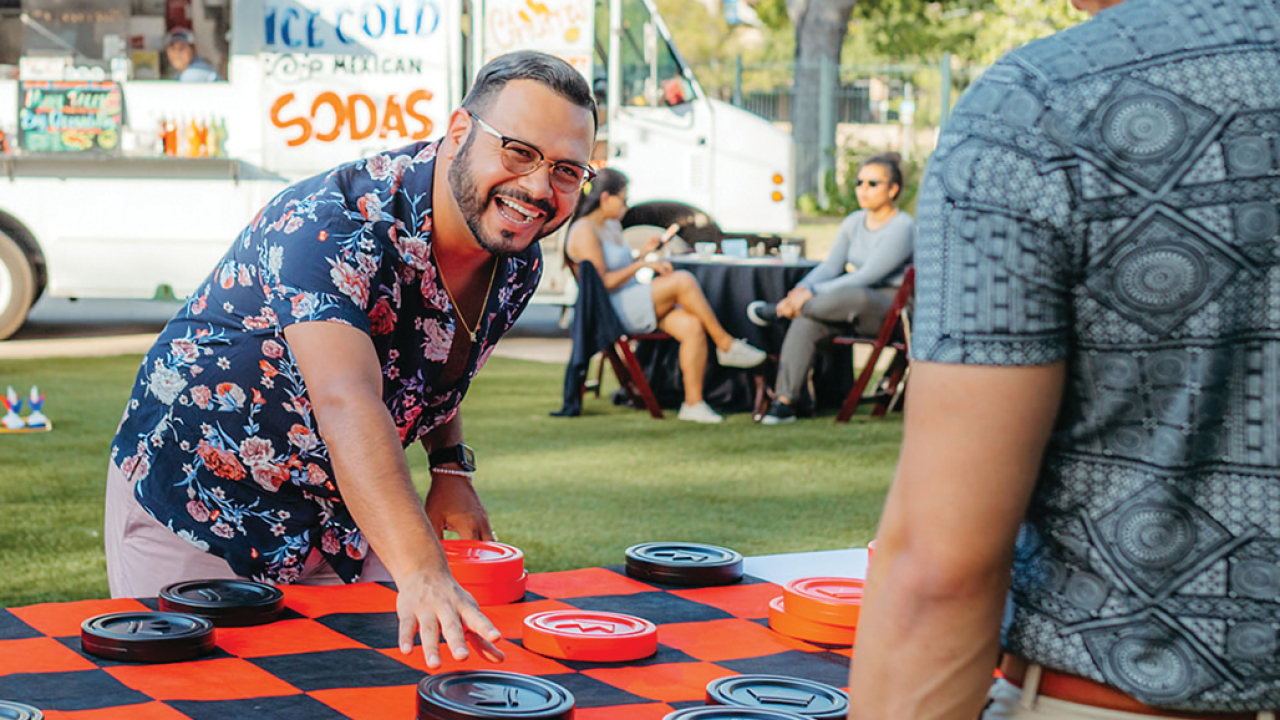 Two people playing a giant game of checkers on a lawn with a food truck in the background.
