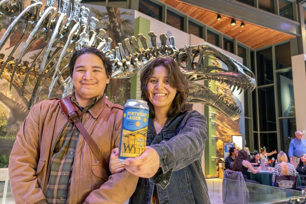 Two young adults in a museum space with a dinosaur skeleton in the background, holding up a can of beer.