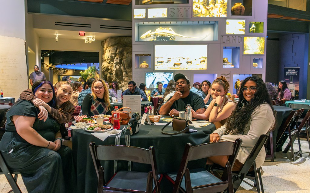 Group of young adults sitting and eating around a round table inside a museum space.