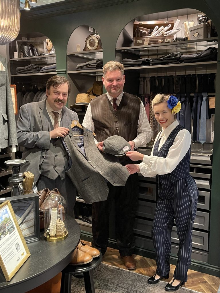 Two men and a woman are standing in a 1930s clothing store and displaying a vest and hat.