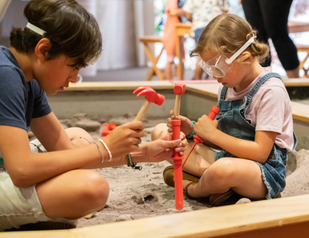 Two children wearing goggles in a sandbox, playing with chisels and  hammers. 