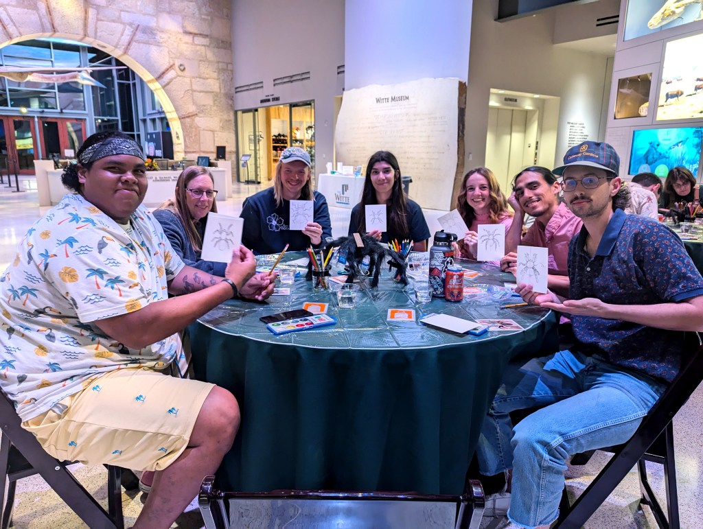 Group of young adults sitting around a round table holding up drawings of a spider inside a museum space.