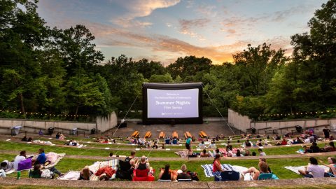 People sitting on outdoor bench seating in front of a screen that says "Welcome to The National Bank of Indianapolis Summer Nights Film Series" with the Newfields logo underneath