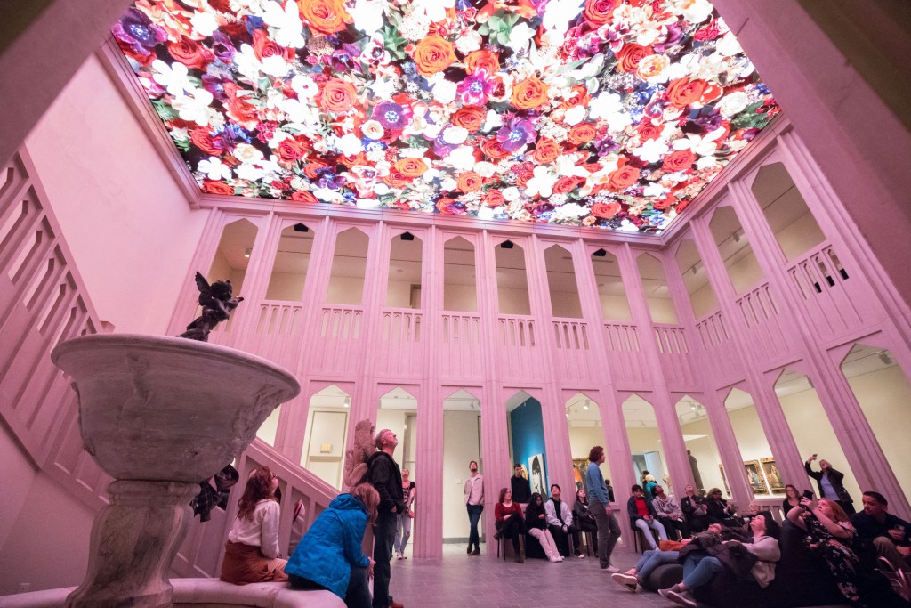 People in a museum atrium looking up at a digital project of flowers on the ceiling