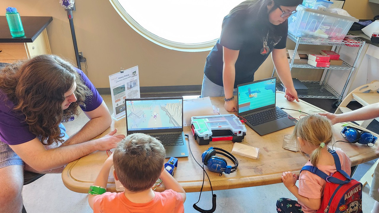 Group of children sitting at a desk, playing games on a laptop with two adults instructing them.
