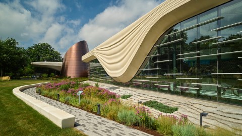 A very modern looking glass building with a walkway of grey bricks and plants in front of it.