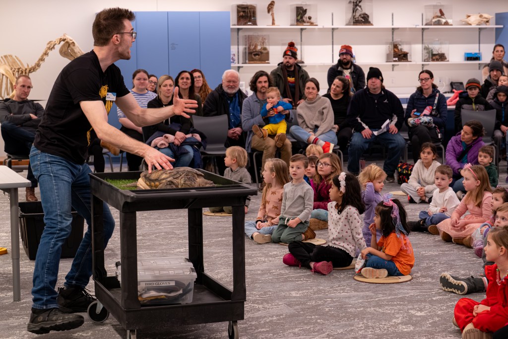 A man is talking to a group of young children, standing in front of a cart with a live turtle on it.