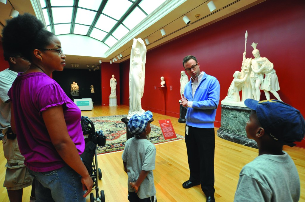 Museum docent interacting with a family with two children in a museum gallery with white sculptures and red walls.