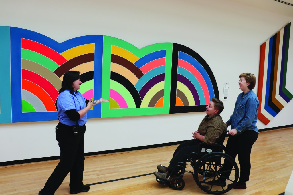 Two visitors, one standing and one in a wheelchair, talk to a gallery guard in front of a brightly colored artwork