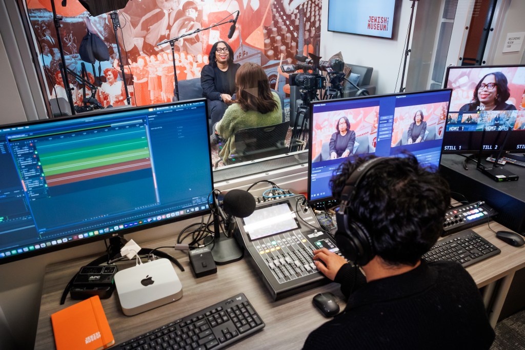 Man behind two computer screens and several keyboards, recording and editing a video of two people talking.