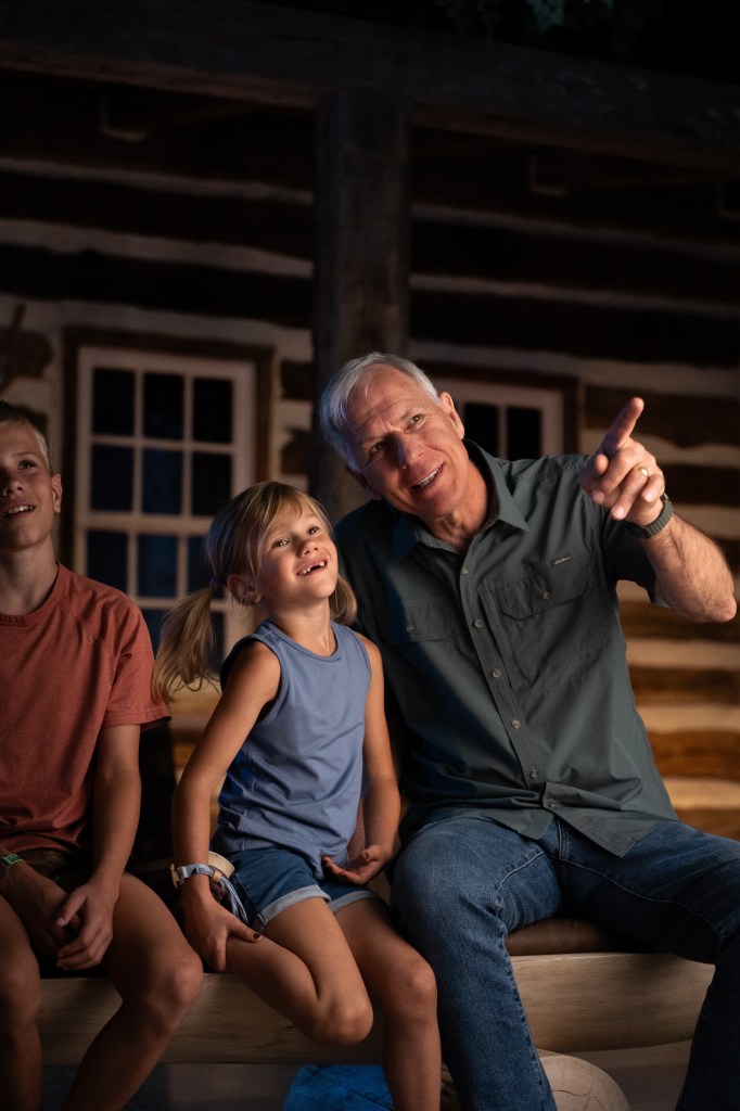 Man with gray hair explaining something to a young girl, pointing at something out of the picture. They are sitting inside a historic cabin.