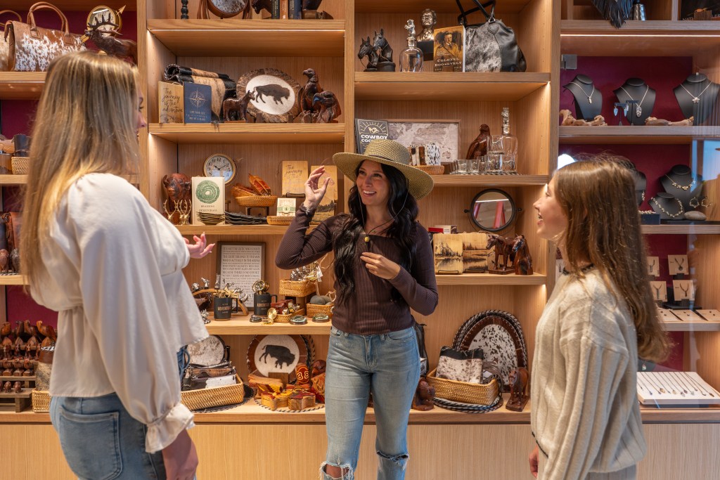 Three women in a museum store, one of them is trying on a hat in front of a merchandise display.
