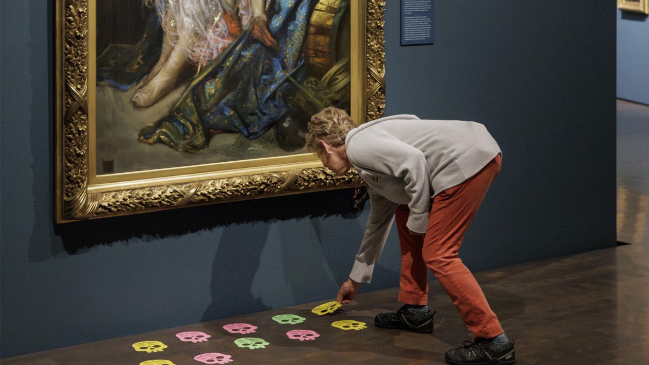 A museum visitor wearing a gray sweatshirt and orange pants bends down to place a two-dimensional yellow paper skull in front of The Family of Street Acrobats: The Injured Child (1873) by Gustave Doré. Ten paper skulls in yellow, pink, and green are scattered in two rows across the dark wooden floor. The gallery walls are a shade of dark blue. Two additional portraits are visible in the background.